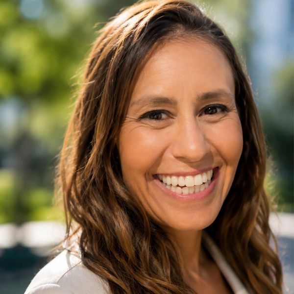 A woman with long brown hair smiling outdoors on a sunny day.