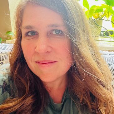 A woman with long wavy hair smiling softly indoors near plants.