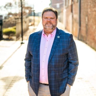 Man in a blue plaid blazer and pink shirt stands outdoors on a sunny day.