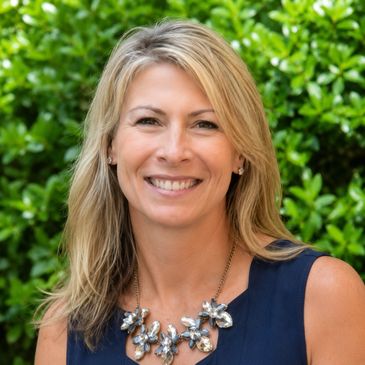 Smiling woman with blonde hair outdoors wearing a navy top and statement necklace.
