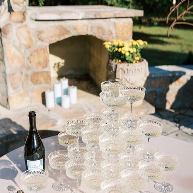 Champagne glasses stacked in a pyramid outdoors with a stone fireplace background.