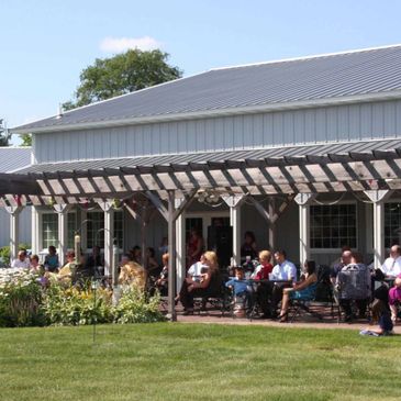 People gathered under a pergola outside a large building on a sunny day.