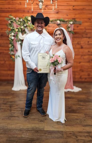 Newlywed couple smiling, holding marriage certificate, dressed in wedding attire.