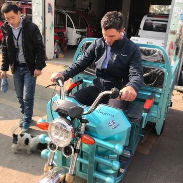 Man testing a turquoise three-wheeled vehicle while another man and a dog stand nearby.