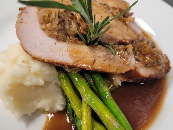Sliced stuffed pork loin with asparagus, mashed potatoes, and gravy, garnished with rosemary.