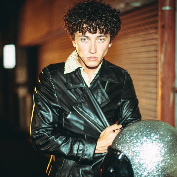 Young man with curly hair wearing a black leather jacket holding a sparkly helmet.