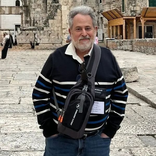 Man with gray hair and beard standing on a stone-paved street.