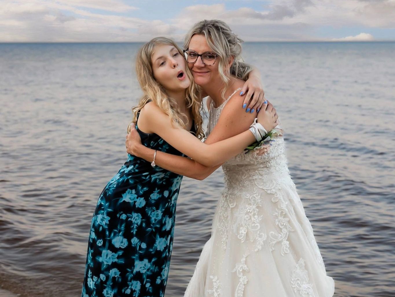 Two women embracing happily by the ocean, one in a wedding dress.