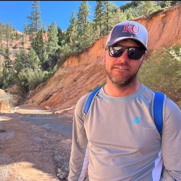 Man wearing sunglasses and a KU cap hiking in a sunny, rocky outdoor area.