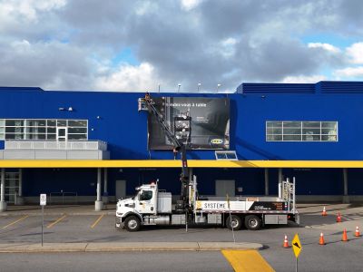 Un camion avec une grue installe une grande affiche sur un bâtiment bleu.