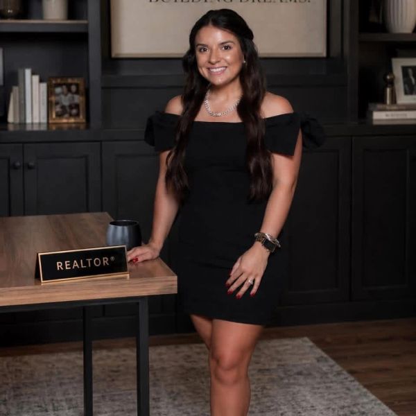 A smiling woman in a black dress stands by a desk with a REALTOR nameplate.