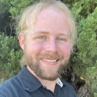 Smiling man with blonde hair and beard outdoors near greenery.