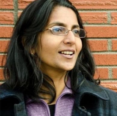 Woman with glasses smiling against a brick wall background.