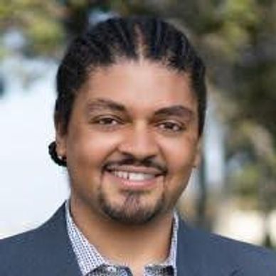 Smiling man with braided hair in a suit jacket outdoors.