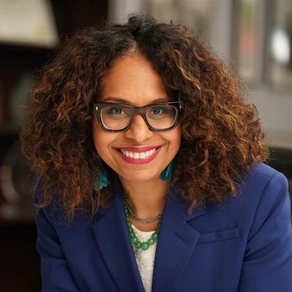 Smiling woman with curly hair and glasses in a blue blazer.