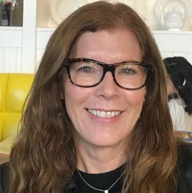 Smiling woman with long brown hair and glasses sitting indoors.