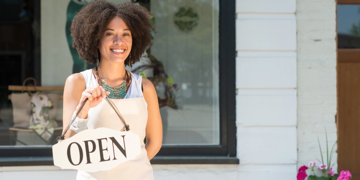 Smiling woman holding an open sign outside a shop.