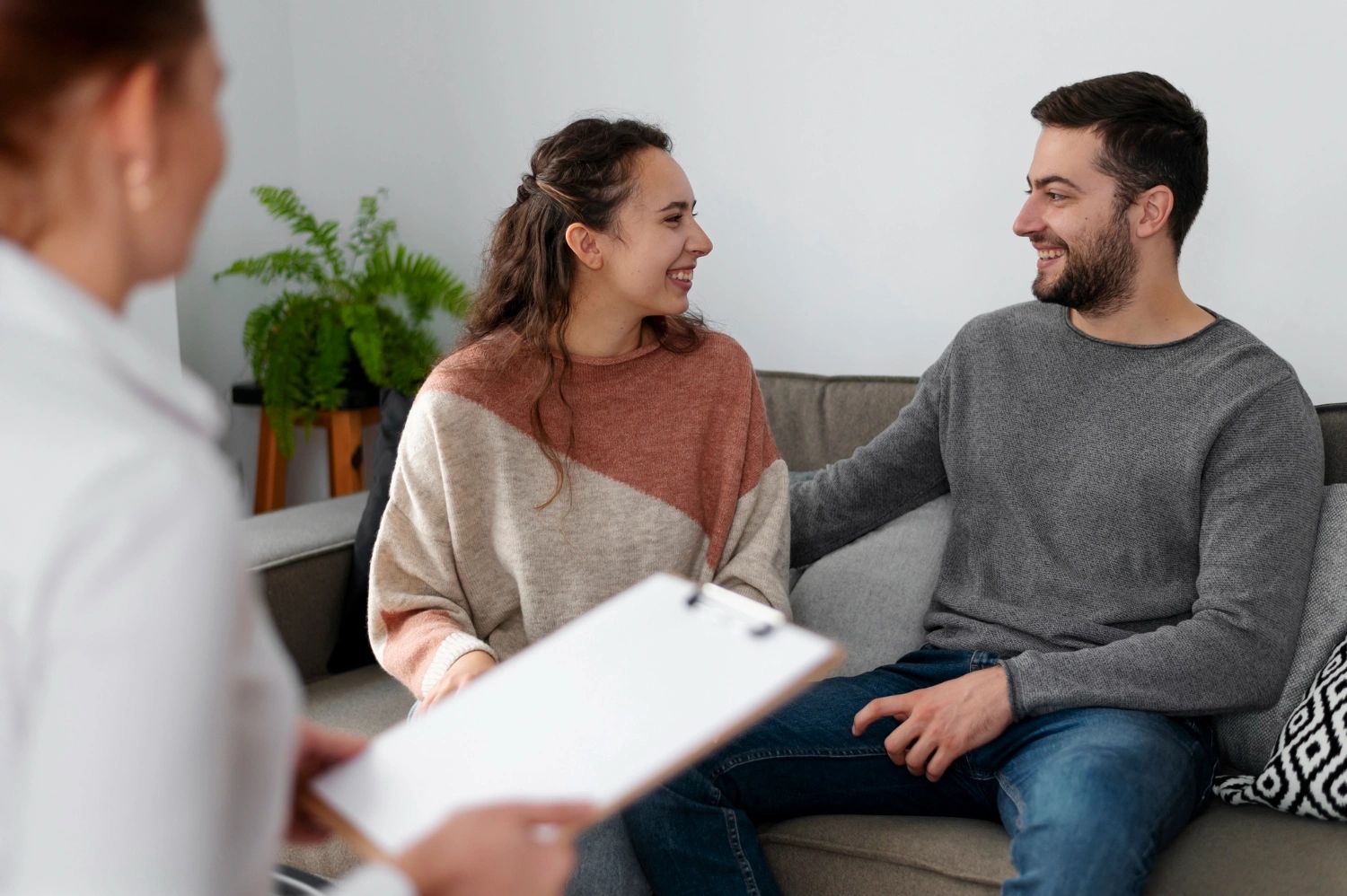 A couple is sitting on a couch, smiling and engaged in a conversation with a therapist.
