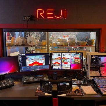 A multimedia control room with multiple monitors and a REJI neon sign.