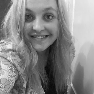 Image of,female presenting adult smiling with lilac hair and a floral orange and yellow top