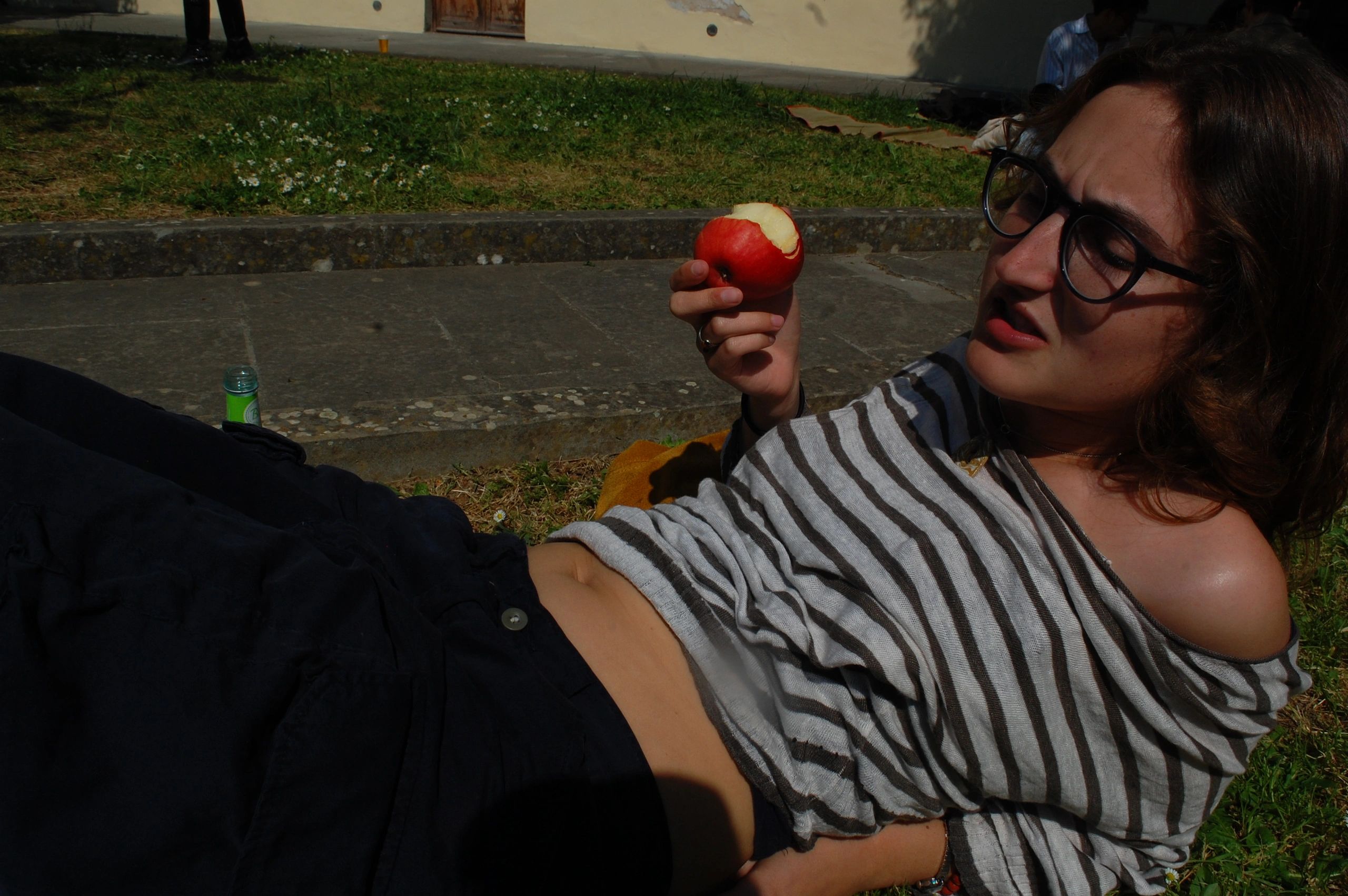 Young woman lying on grass, holding a bitten red apple, wearing glasses and a striped shirt.