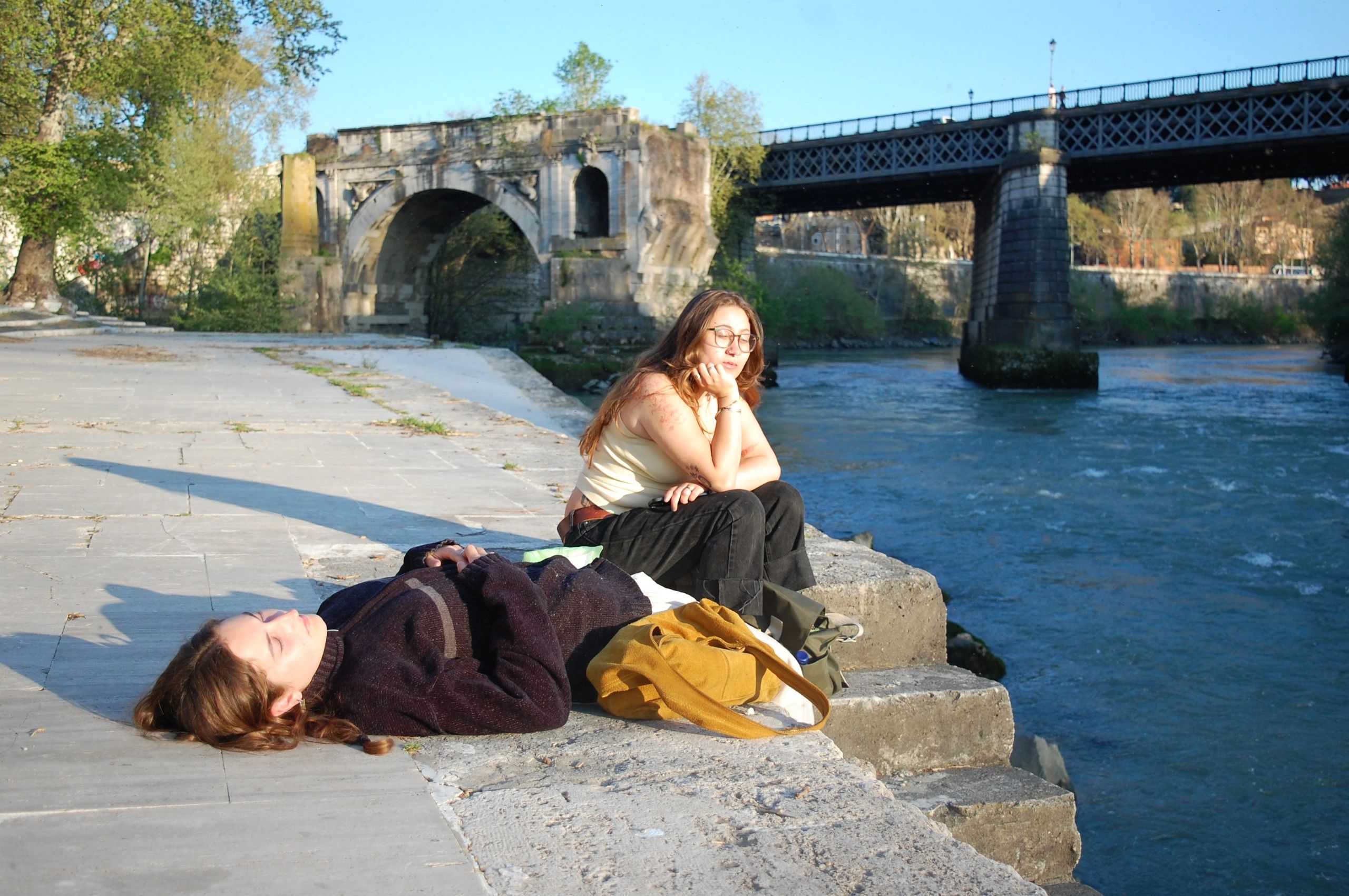 Two women relaxing by a river near an old stone bridge on a sunny day.