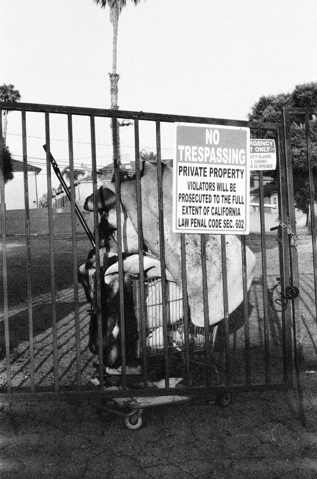 Black and white image of a gate with a no trespassing sign and a shopping cart full of items behind it.