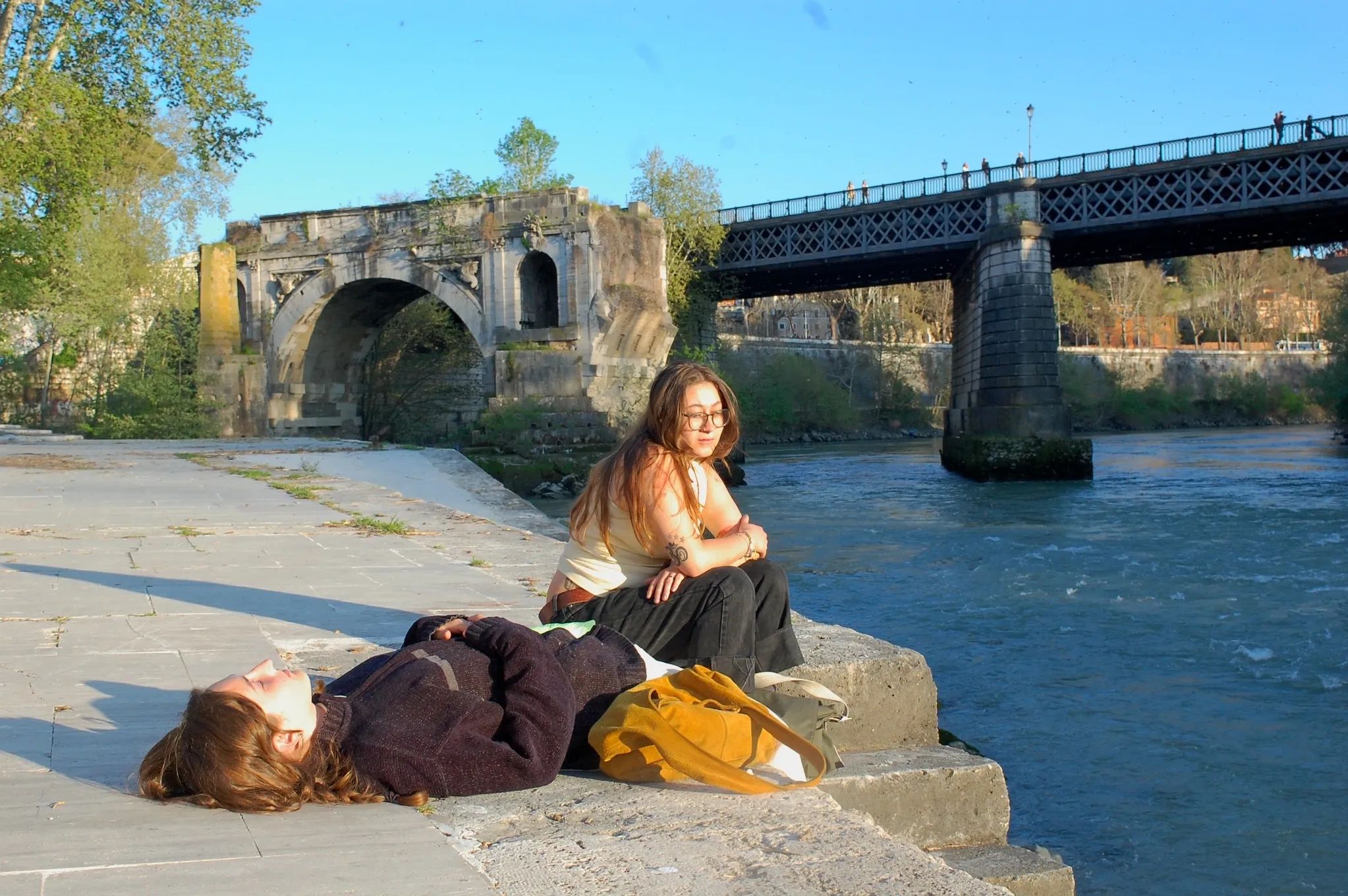 Two people relaxing by a river near historic bridges on a sunny day.