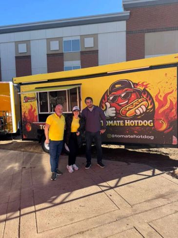 Three people posing in front of a bright yellow tomato hotdog food truck.
