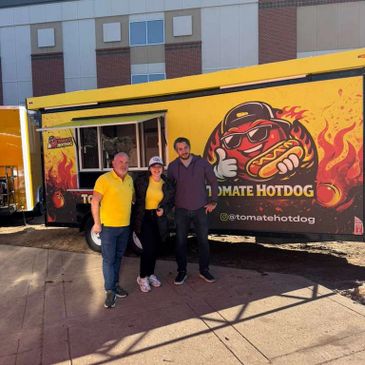 Three people posing in front of a bright yellow tomato hotdog food truck.