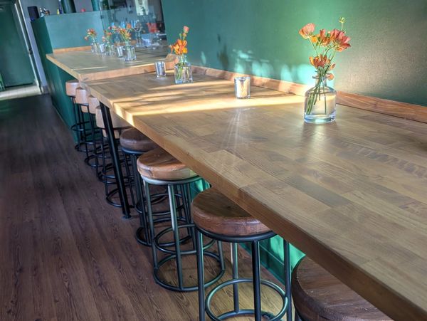 Sunlit wooden counter with stools and small floral arrangements along a green wall.