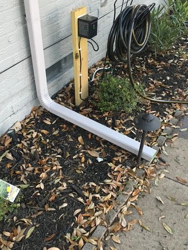 Outdoor garden area with a downspout, hose, and scattered leaves.
