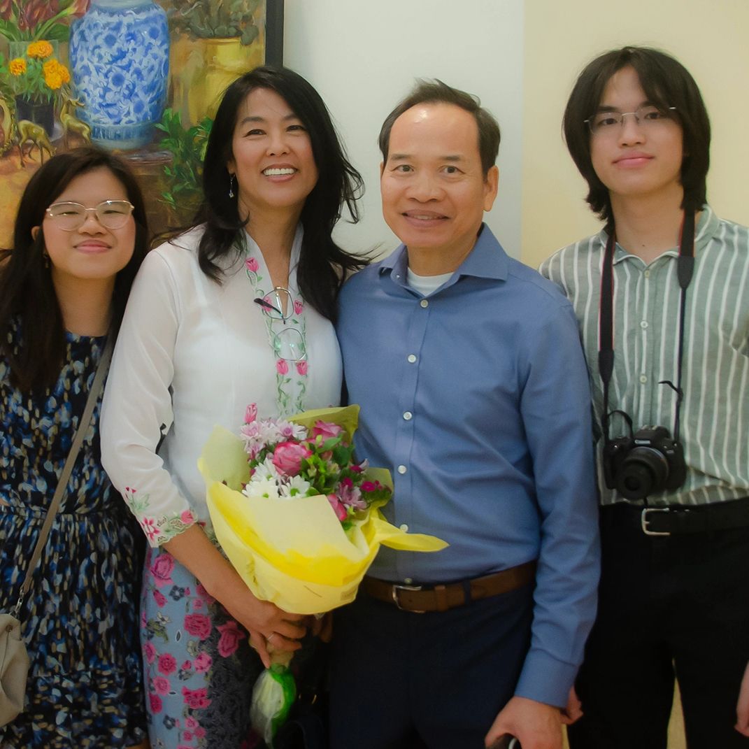 A smiling family of four posing indoors, with the mother holding a bouquet of flowers.