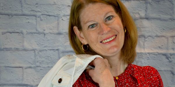 Smiling woman with red hair holding a white jacket over her shoulder against a brick wall.