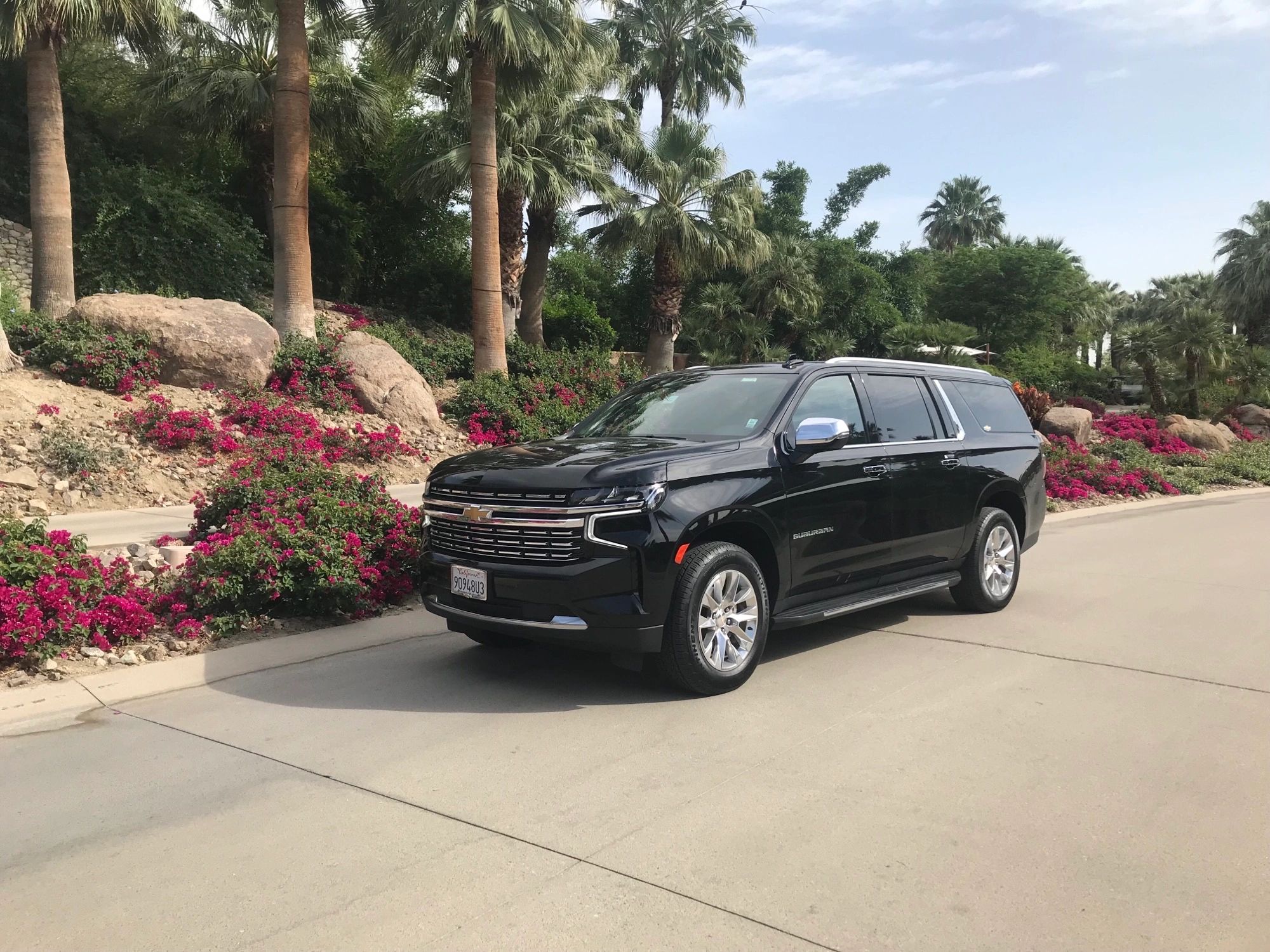 Black Chevrolet Suburban parked on a sunny street with palm trees and flowers.
