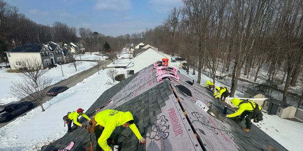 Workers in bright yellow jackets installing roof shingles on a snowy day.