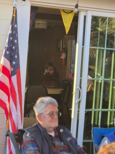 An elderly man sitting outside near an American flag and a glass door.