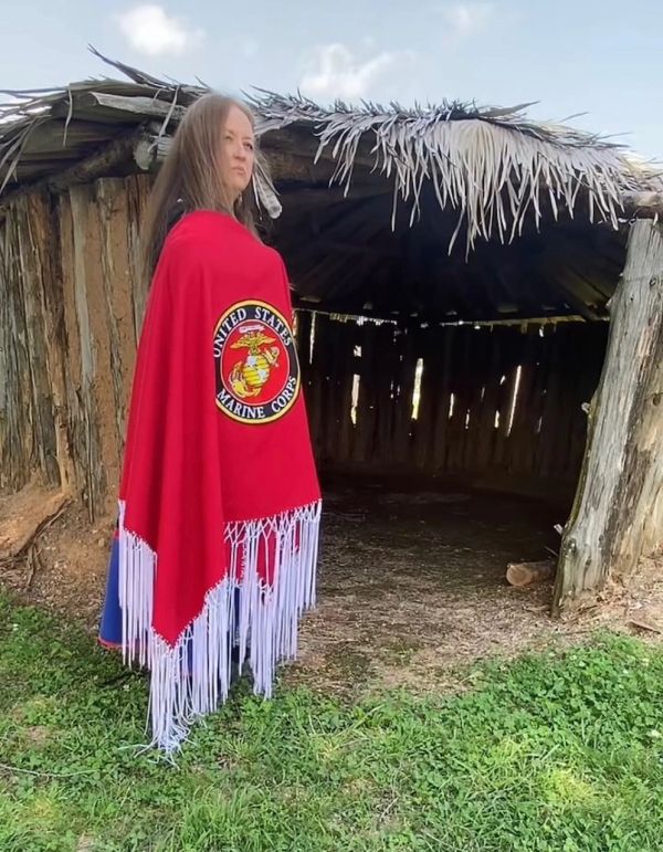 Person wearing a red United States Marine Corps shawl near a rustic hut.