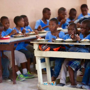 Children in blue shirts eating together at tables in a communal setting.