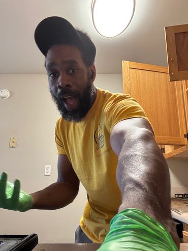 Man in yellow shirt and green gloves reaches toward the camera in a kitchen.