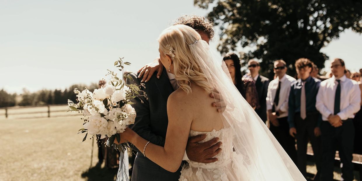 Bride and groom embrace outdoors with guests in the background.