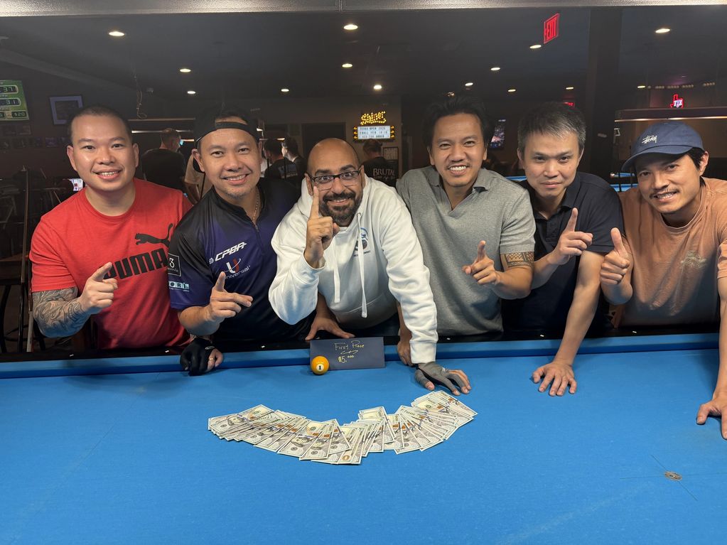 Six men celebrating a pool game win with money and an 8-ball on the table.