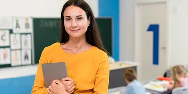 Female teacher holding a tablet in a classroom with young students.