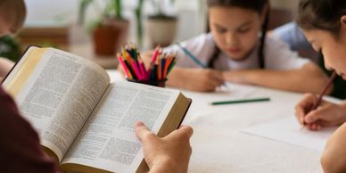 Person reading a book while children write at a table.