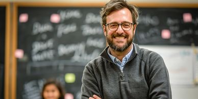 Smiling male teacher standing in front of a classroom blackboard.