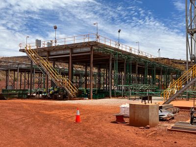 Industrial site with metal structures and orange dirt ground under a partly cloudy sky.