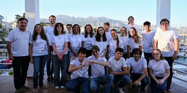 Group of young people and adults posing outdoors near a marina, wearing matching white shirts.