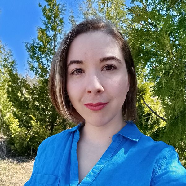 Young woman in a blue shirt smiling outdoors with greenery and blue sky.