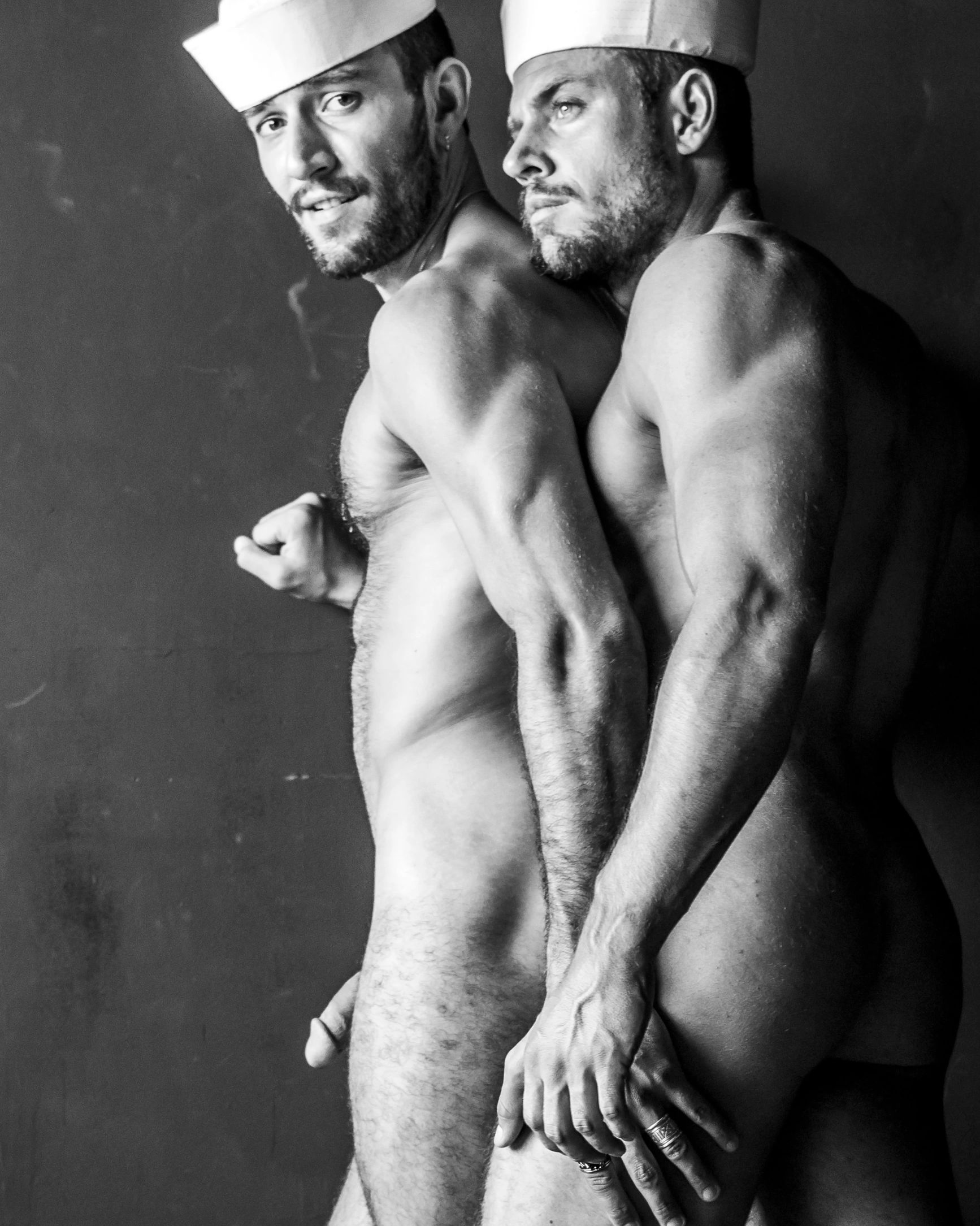 Black and white photo of two shirtless men wearing sailor hats, posed closely together.