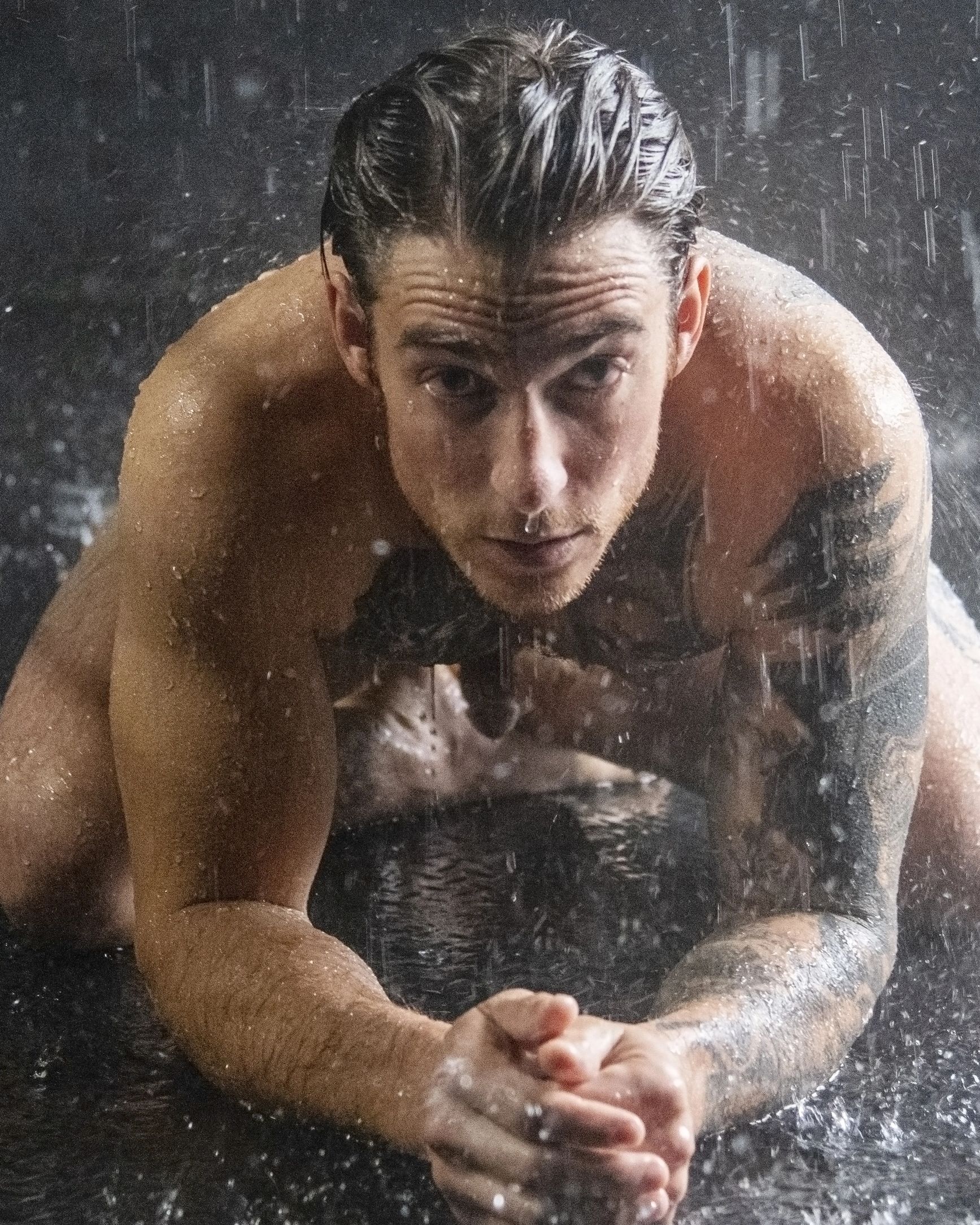 Man with wet hair and tattoos under water droplets, looking intensely ahead.